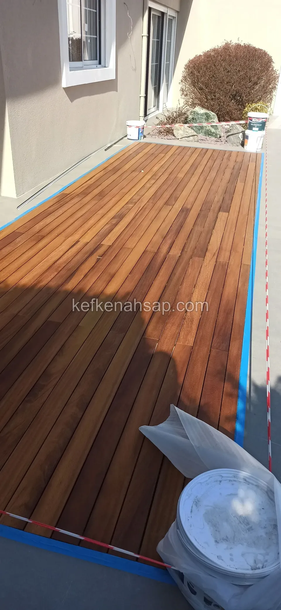 Ev duvarı boyunca uzanan, kenarları mavi bantla korunmuş koyu turuncu deck ve önde beyaz kovalar