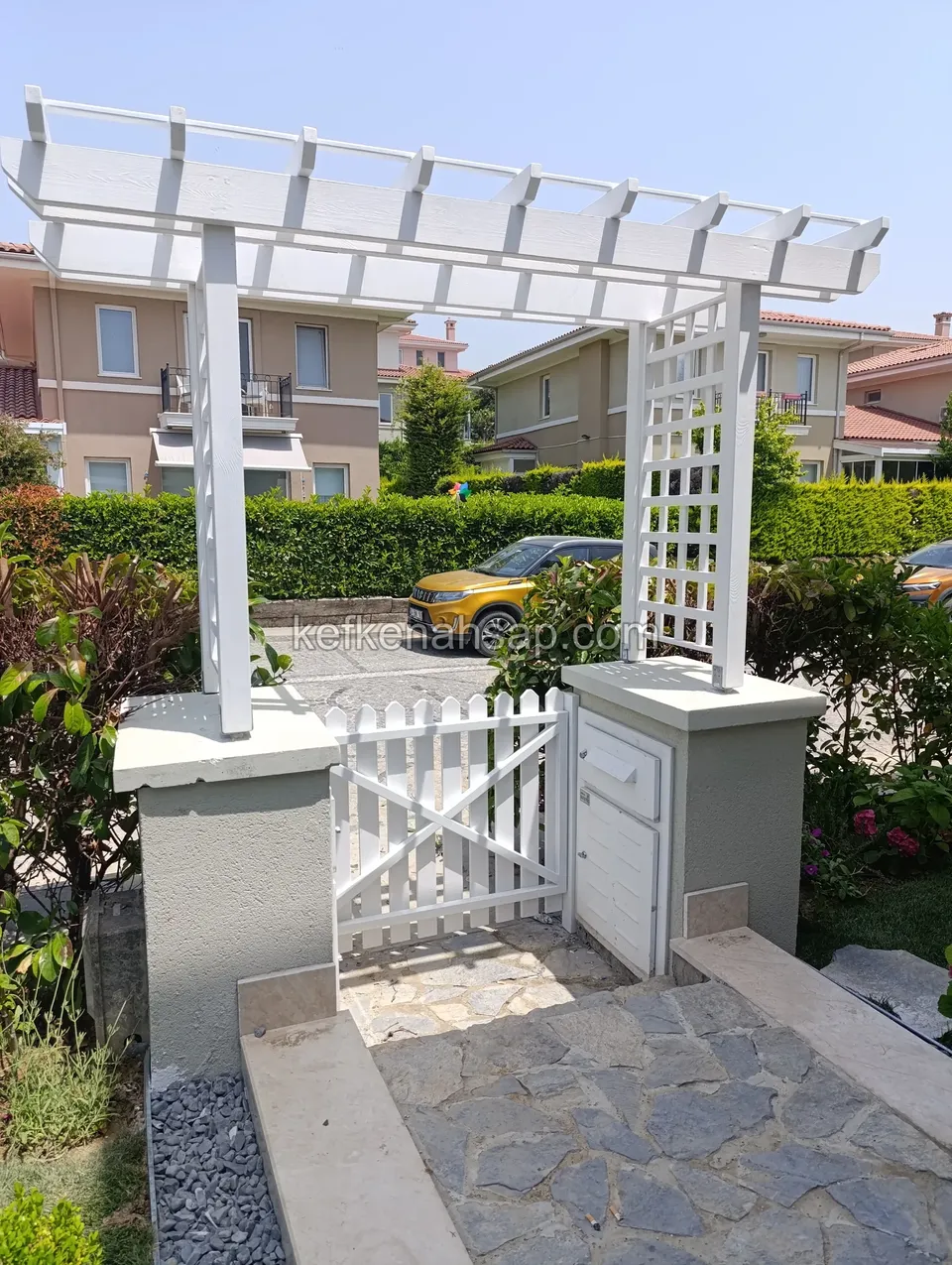 Beyaz boyalı pergola kemerinin altında çıtalı bahçe kapısı, kapı direkleri, taş basamaklar ve taş döşeli yol