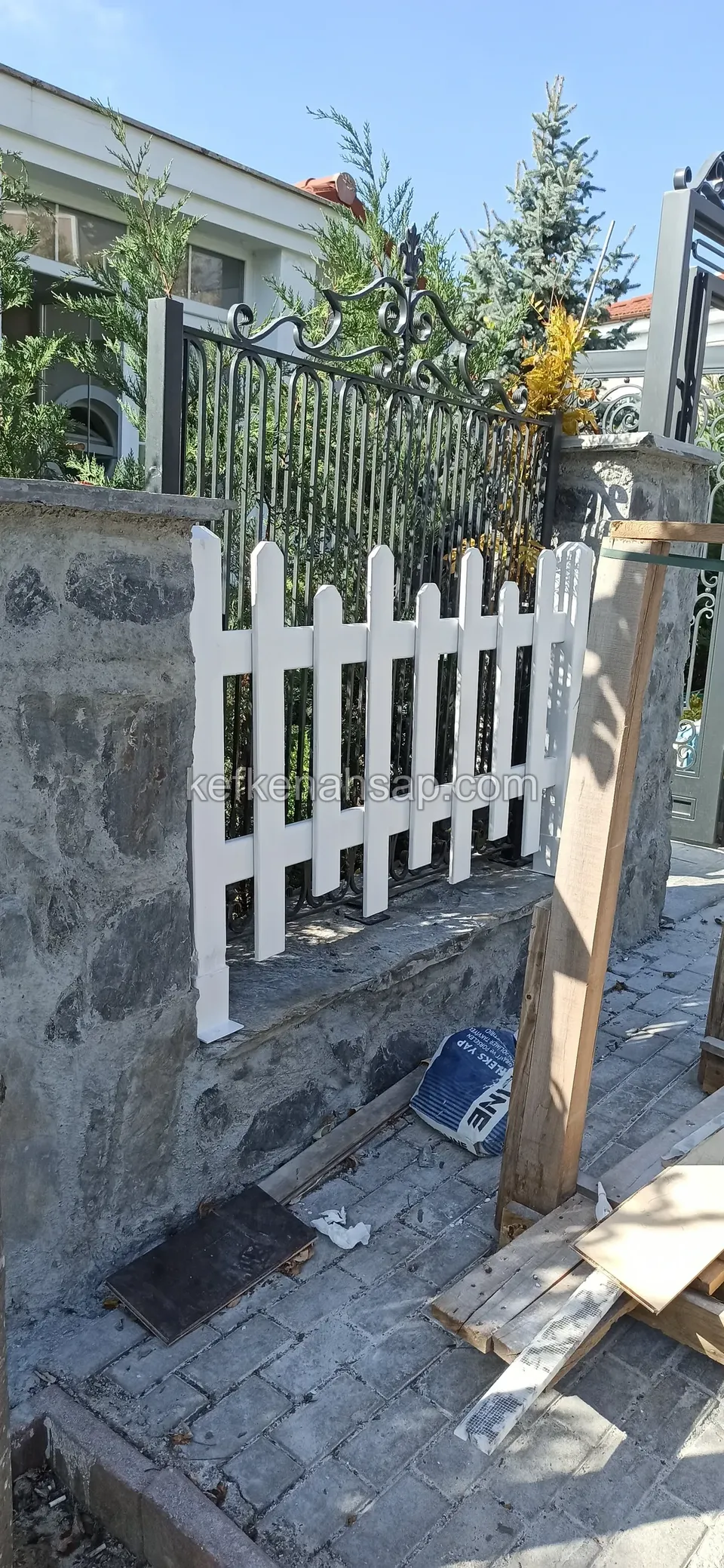 Taş duvar üstüne takılmış beyaz çıtalı çit, yanında dikili bir ahşap direk, yerde tahta parçaları ve alet kutusu