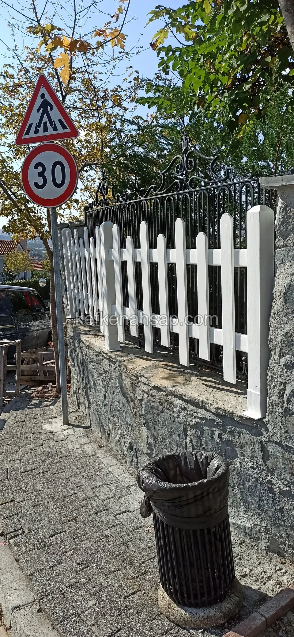 Taş duvar üzerinde beyaz boyalı çıtalı çit, yanında yaya geçidi ve hız sınırı levhası, önde çöp kovası