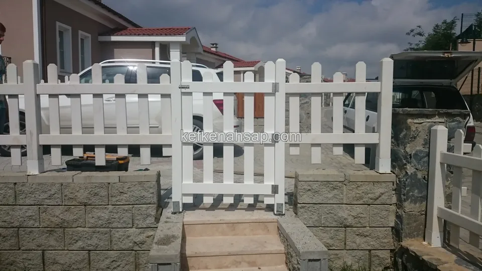 Taş kaplı basamaklı girişte beyaz boyalı çıtalı kapı ve iki yanda çit, arkada park edilmiş beyaz bir araç ve evler