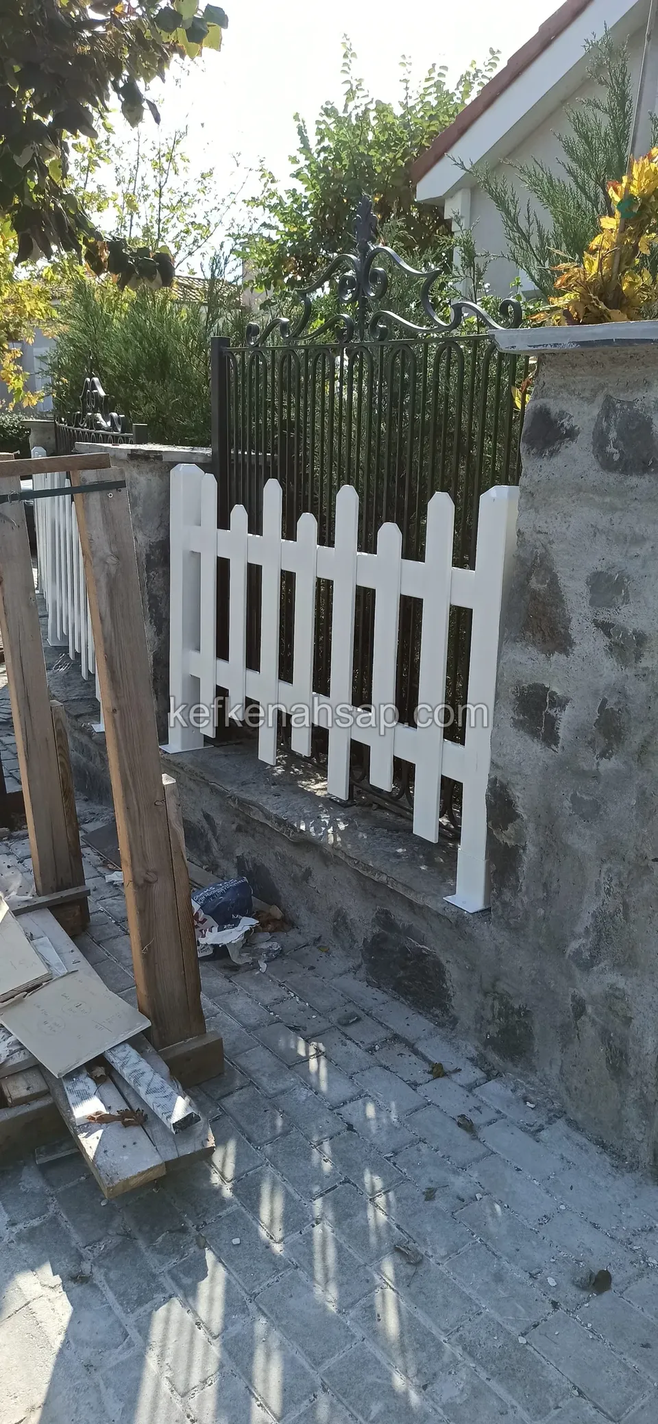 Taş duvarın üzerindeki beyaz çıtalı çit, solda dikili ahşap kirişler ve yerde tahta yığını