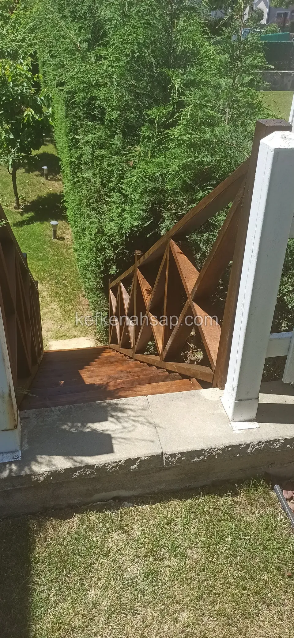 Beyaz bir sütunun yanında, ahşap basamaklara inen kahverengi cilalı çaprazlı korkuluk, arkada yeşil çit ve çimlik