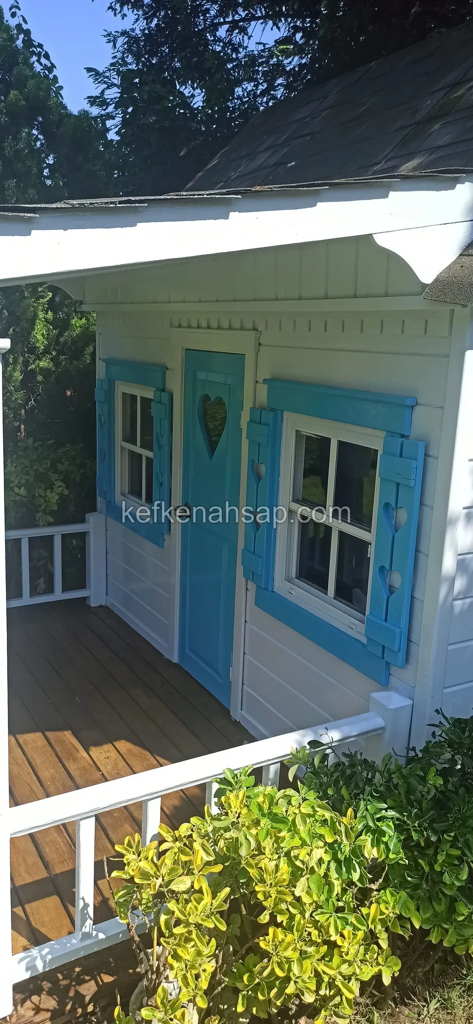 Beyaz boyalı, mavi kapı ve pencere kepenkli, ahşap verandası olan çocuk oyun evi
