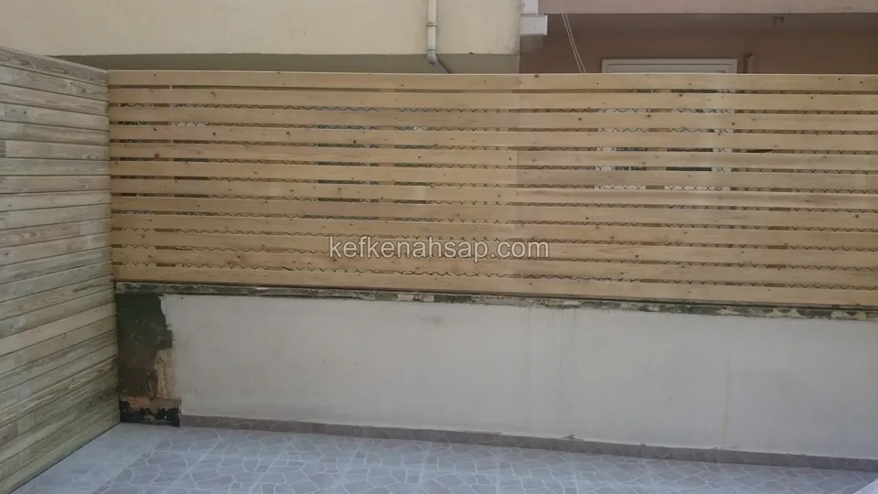 Teras köşesinde bina duvarının önünde uzanan sık yatay çıtalı açık renk ahşap panel; solda dikey tahtalı yan yüz