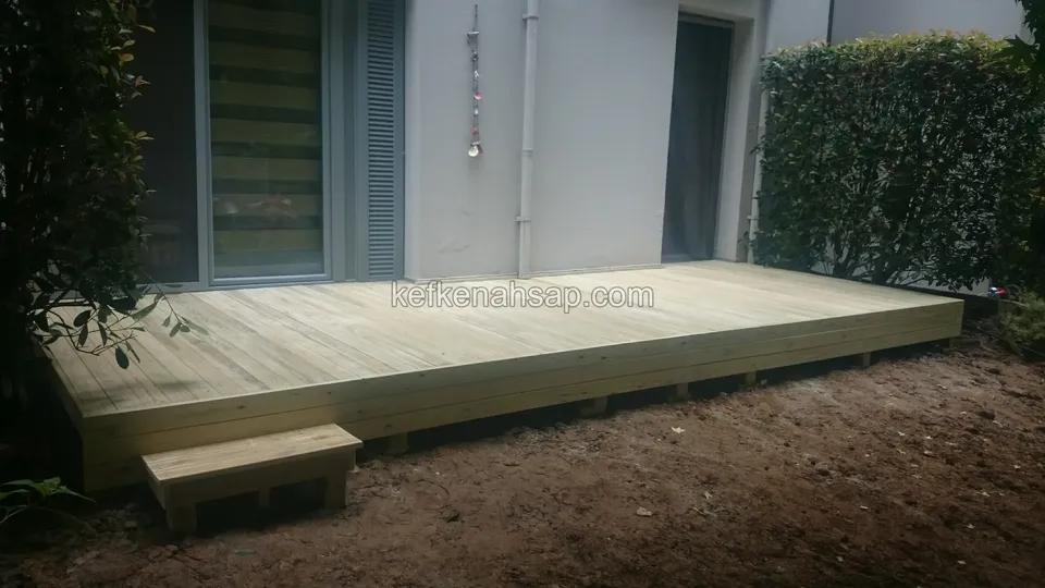 Low timber deck raised slightly above the soil in front of a building's glass door, with a small step at the edge