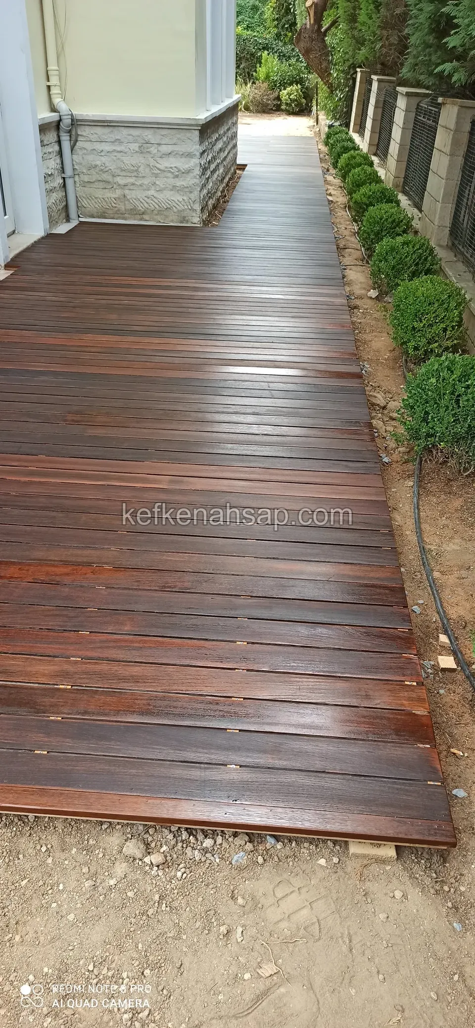 Bina kenarında bitkilerle sınırlanan dar koyu kahverengi deck yürüyüş yolu