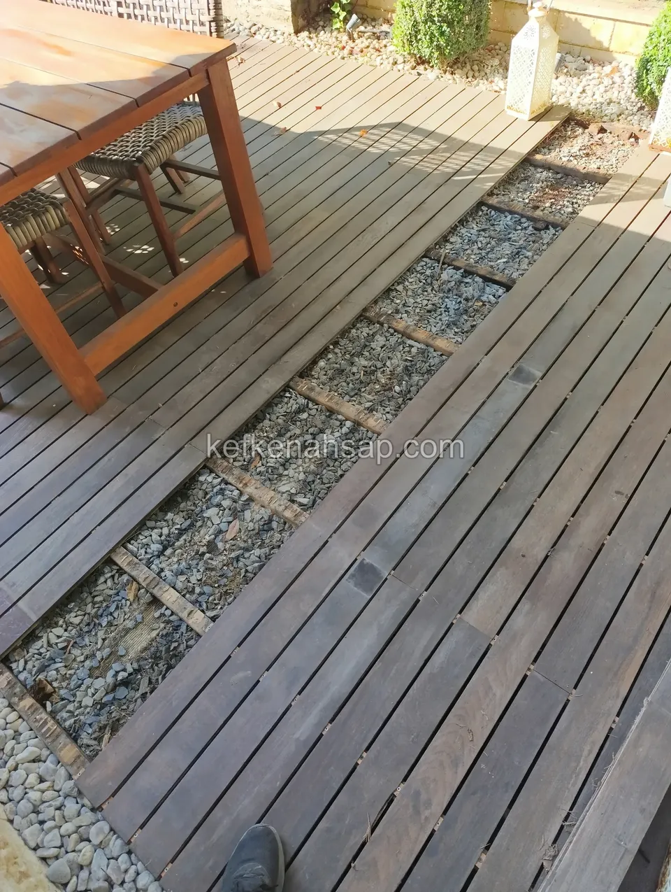 Greyed deck whose boards are separated by gravel strips, with a timber table on the left and the tip of a shoe in the corner