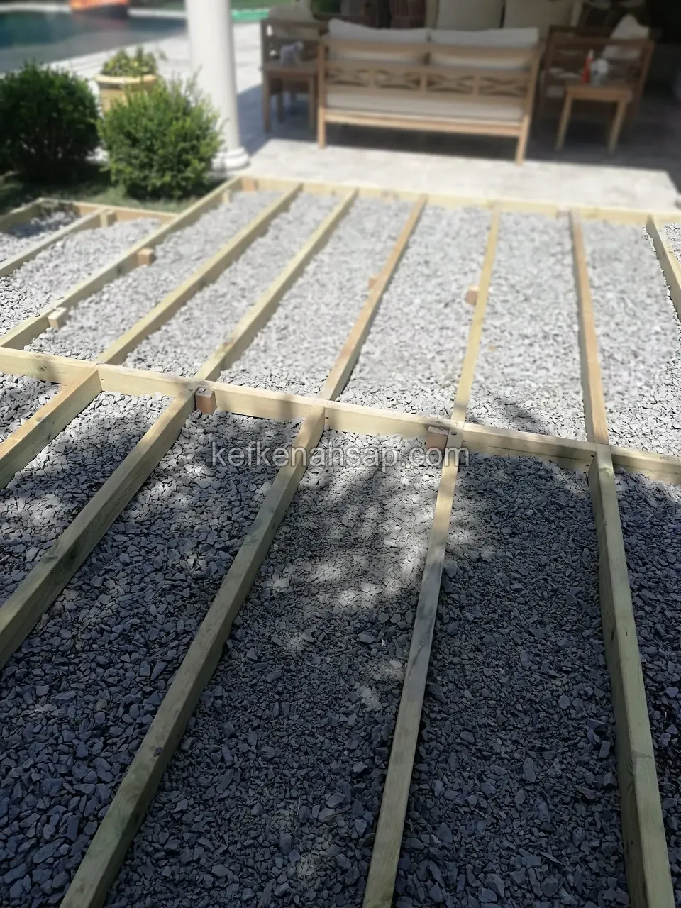 Koyu çakıl üzerinde çapraz ve boyuna uzanan açık renkli deck kirişleri, arkada ahşap oturma grubu