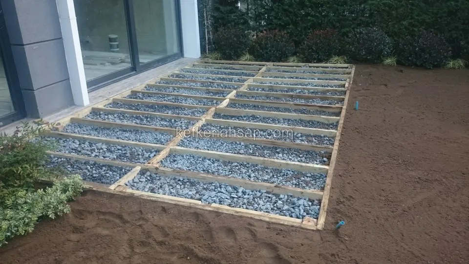 Gravel-filled timber deck substructure with soil and shrubs at its edges and a glass door behind