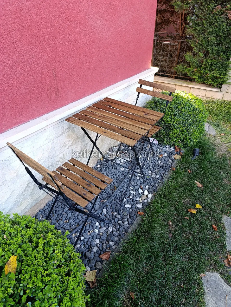 Vor einer rosa gestrichenen Wand stehen auf einem Kieselstreifen zwei Klapp-Gartenstühle aus Holzlatten und ein kleiner Tisch