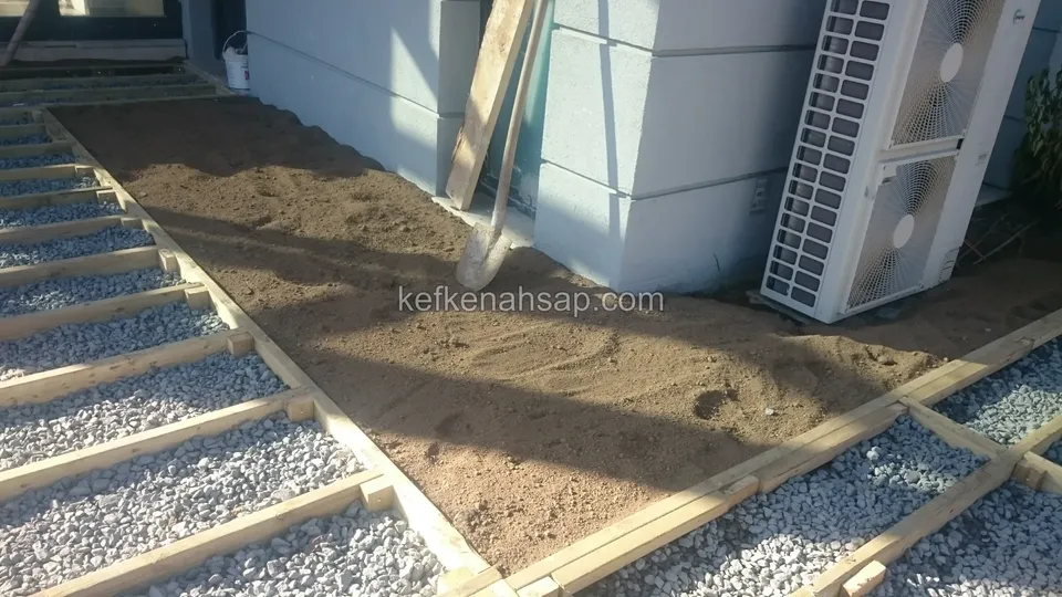 Timber deck substructure on gravel and soil with an air-conditioning unit beside it and a board leaning on the wall