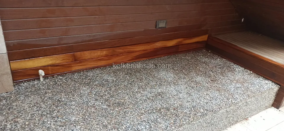 Çakıl zeminli bir alanda yatay ahşap duvar kaplaması ve köşede ahşap oturma bölümü