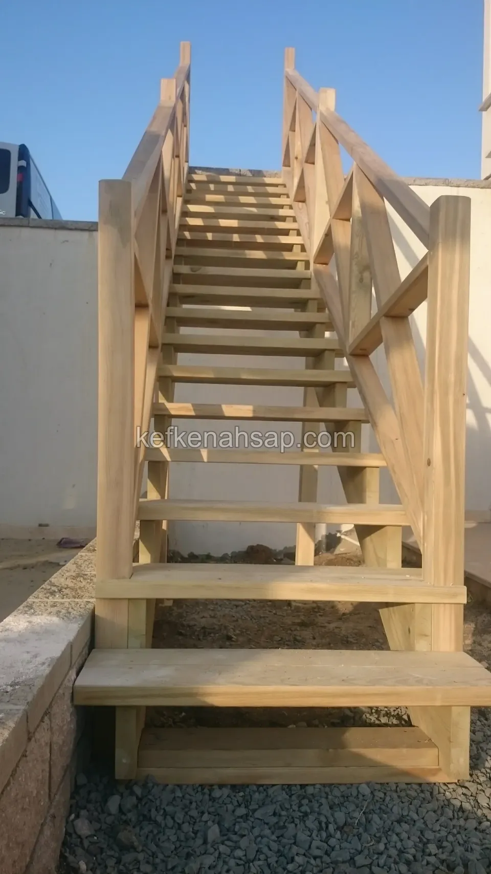 Çakıl döşeli zeminden duvar üstüne çıkan, iki yanı çapraz korkuluklu ahşap merdivenin önden görünüşü