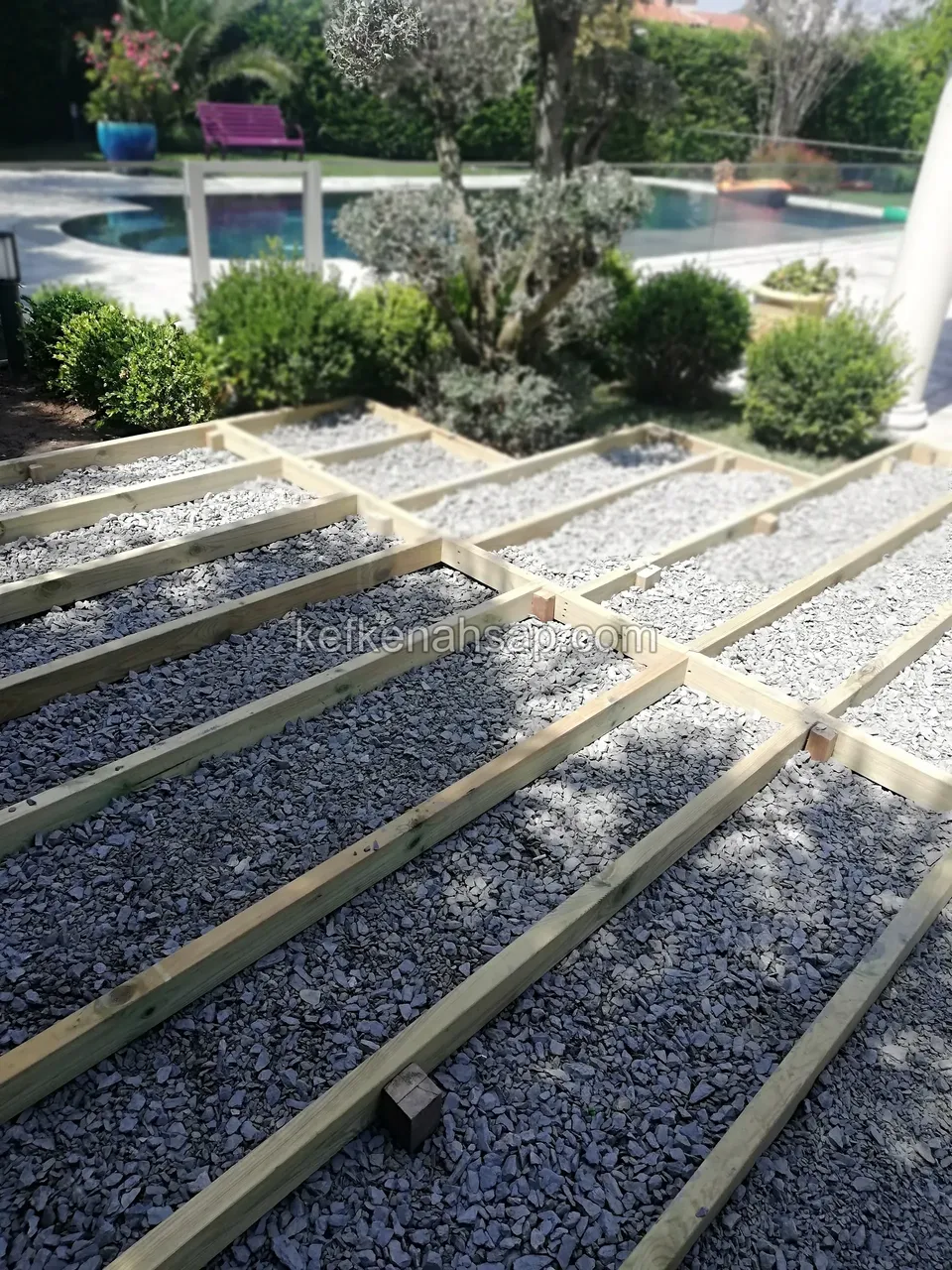 Çakıl üzerine kurulmuş ahşap kiriş çerçevesi, arkada havuz ve bahçe bitkileri