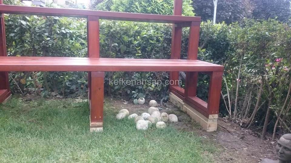 Eine rotbraun gestrichene Holzbank mit hoher waagerechter Lehnenlatte in einem Rasengarten vor Hecken, davor Steine im Gras