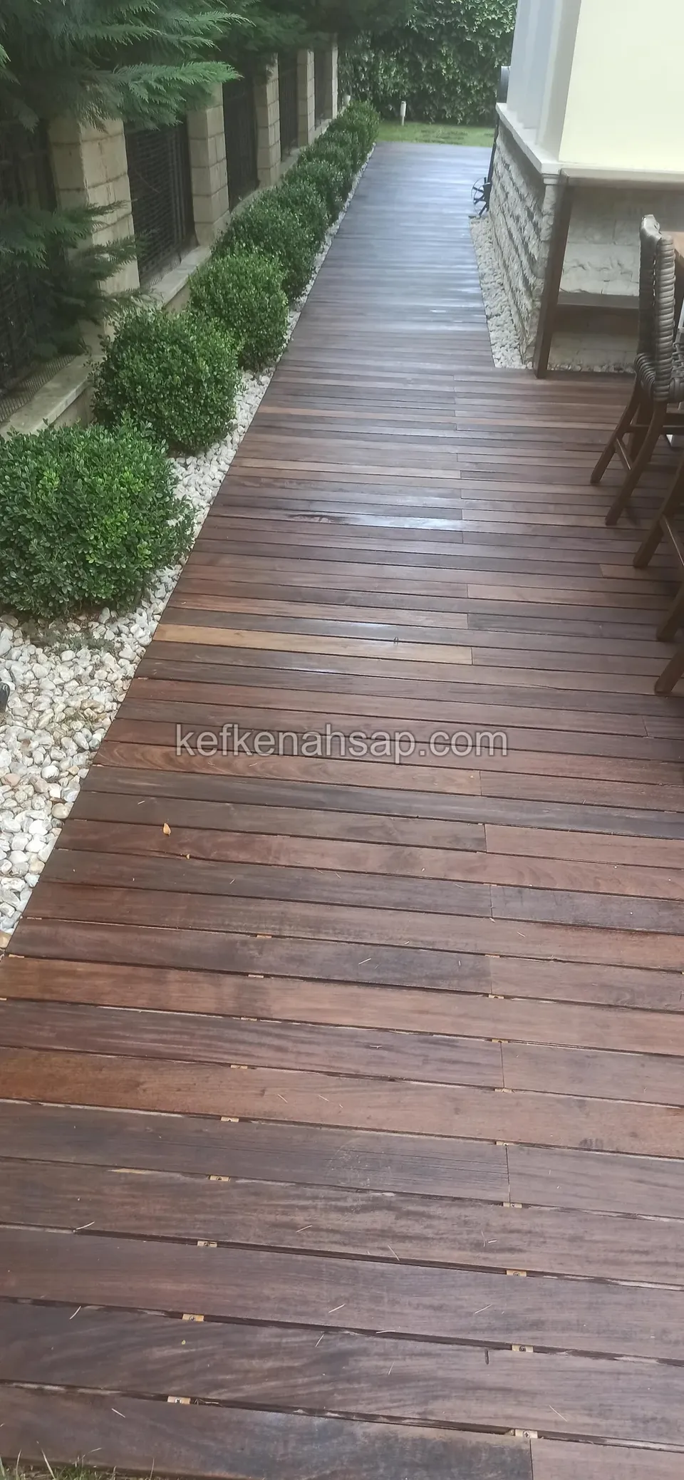 Evin yanında, yuvarlak budanmış çalılar ve beyaz çakıl şeridi boyunca uzanan koyu kahverengi deck yolu