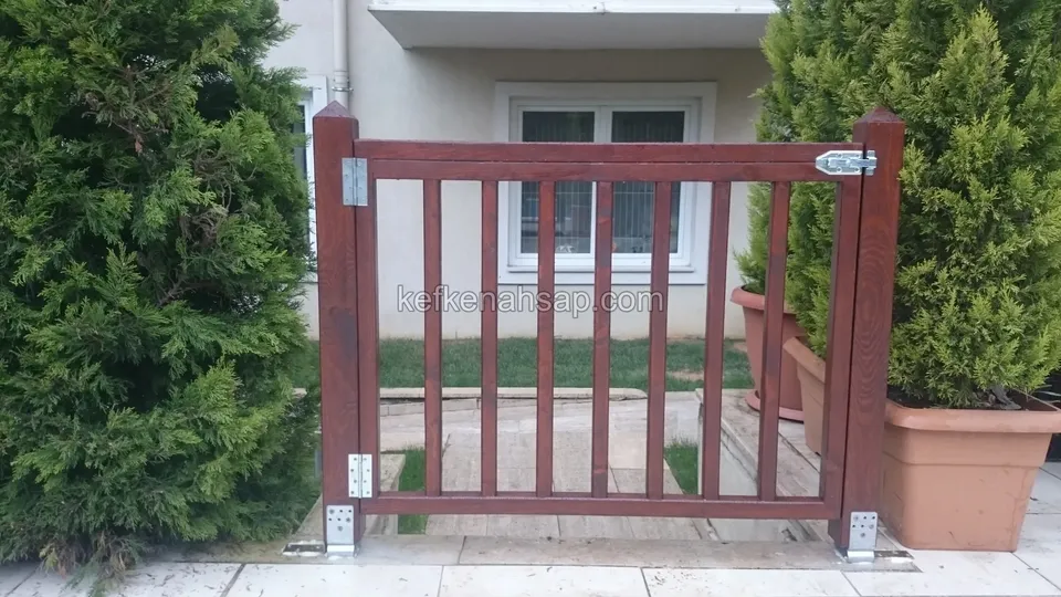 Selvi ve saksı bitkisi arasında duran, dikey çıtalı bordo-kırmızı boyalı küçük ahşap bahçe kapısı; üstte paslanmaz mandal