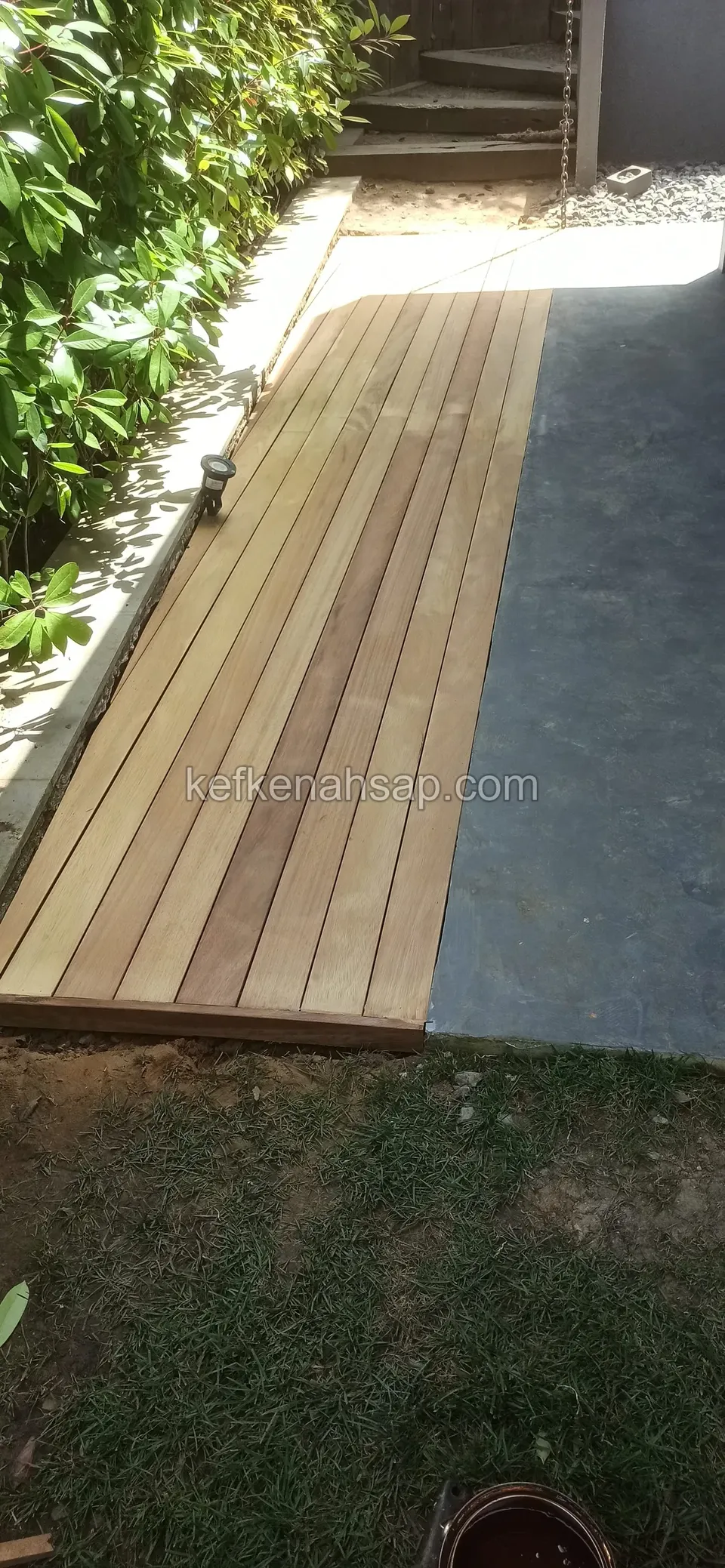 Yan geçitte, çalıların kenarında beton zemine bitişik yeni döşenmiş açık renkli dar deck şeridi