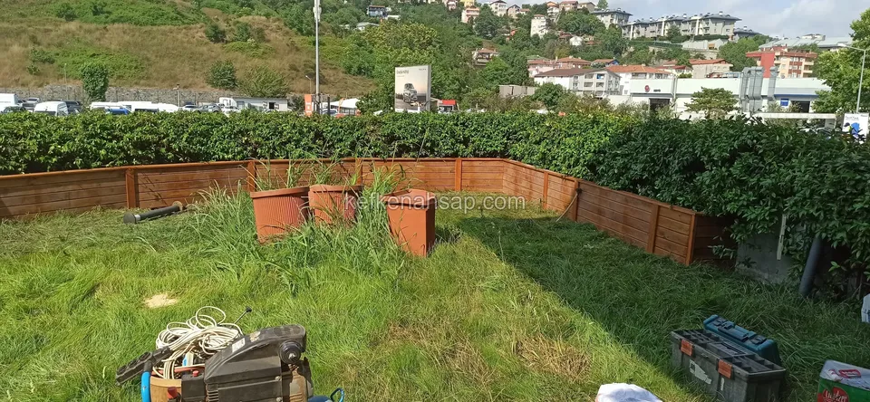 Çalılık önünde çimenin üzerinde köşeli dizilmiş alçak turuncu ahşap çiçeklik kasaları; yerde aletler