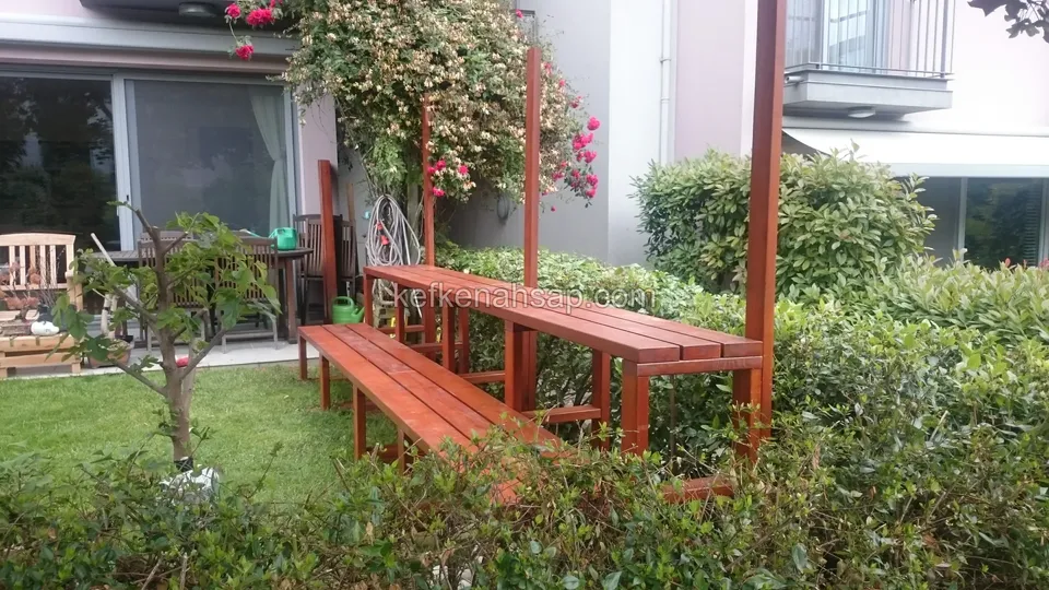 Ein langer rötlich lackierter Holztisch mit Bank in einem Rasengarten zwischen Sträuchern