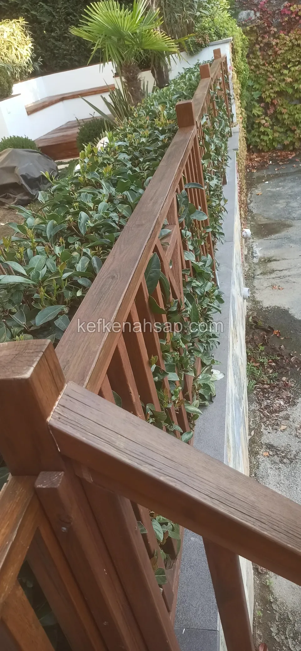 Yakından görünen kahverengi cilalı dikey çıtalı ahşap korkuluk ve küpeşte, yanında yeşil çalılık