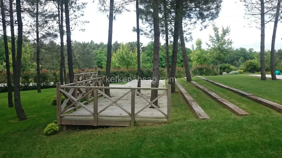 Çimde çam ağaçlarının yanında, çevresi çapraz çıtalı korkuluklu alçak deck platformu; önde çimde uzanan kereste traversler