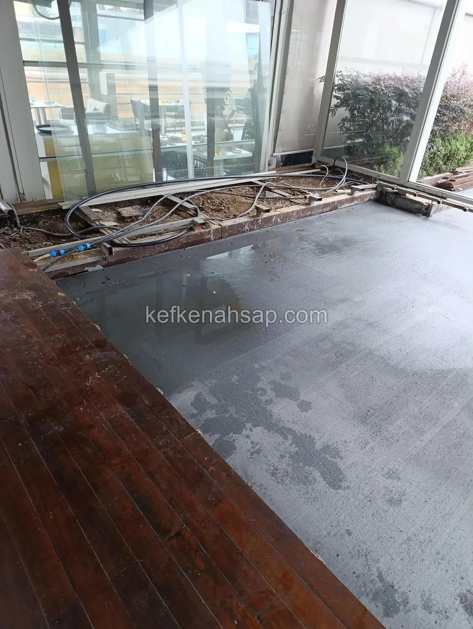 Cam cepheli alanın önünde ıslak gri beton zemin ve sol kenarda koyu kırmızı kahverengi deck; arkada kablolar ve borular