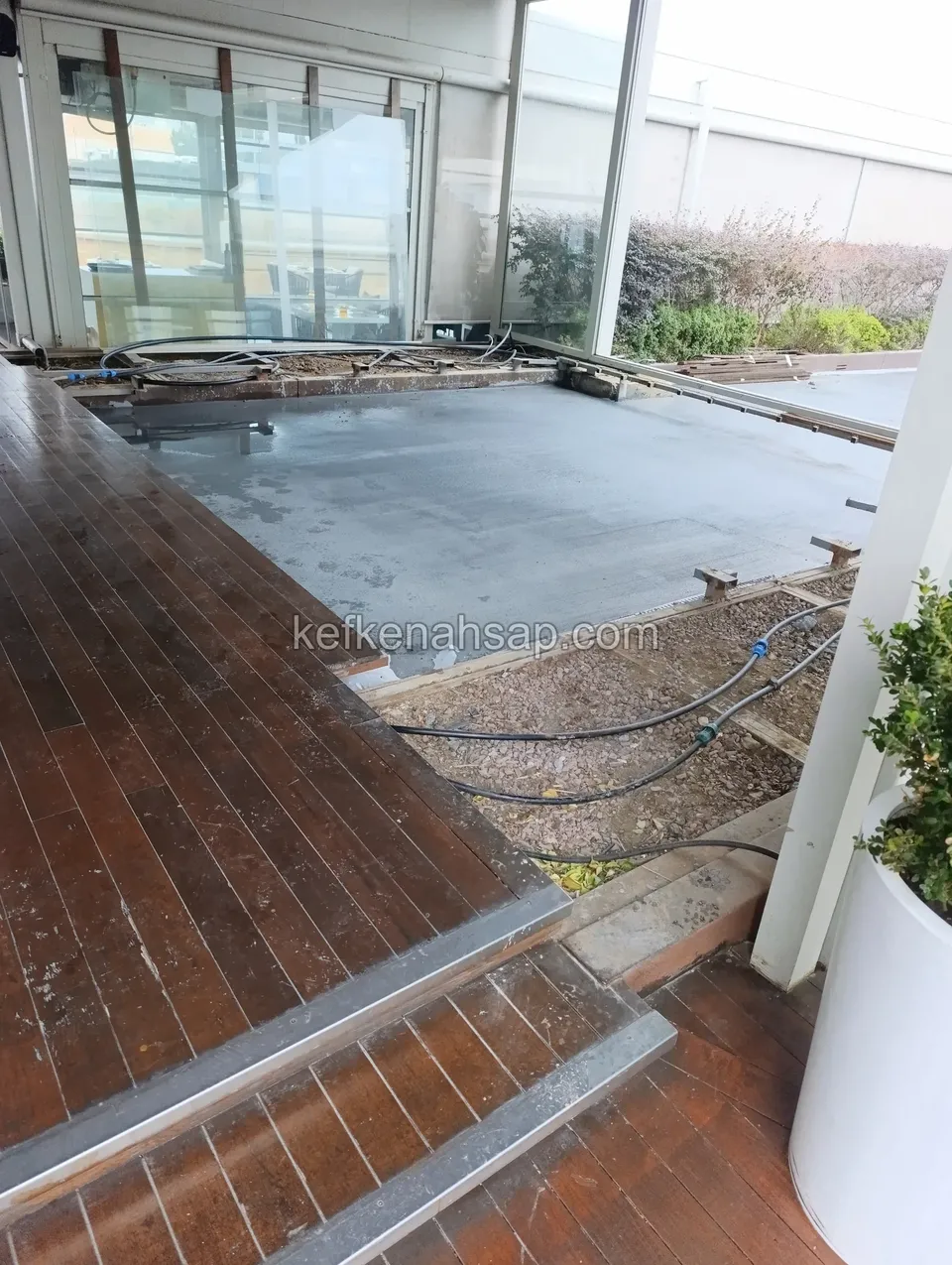 Cam cepheli bir mekânın girişinde koyu deck ve basamaklar; yanında nemli beton zemin ve açıkta kalan borular