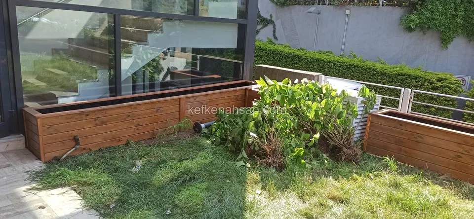Varnished timber planter meeting at a corner in front of a glass wall, with a green plant beside it