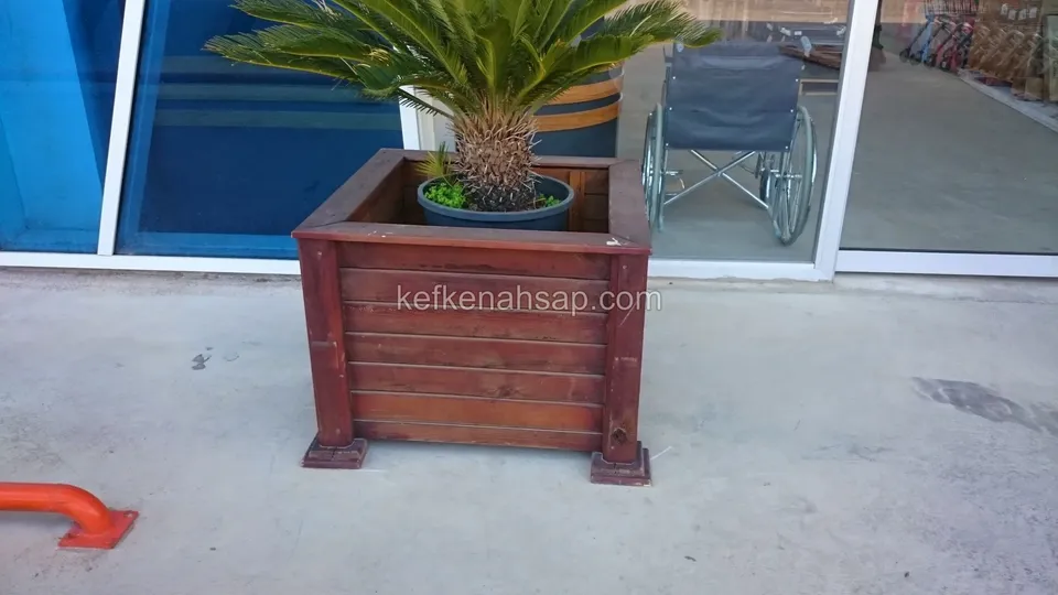 Cam girişin önünde duran, kırmızı-kahve vernikli yatay çıtalı çiçeklik; içinde palmiye