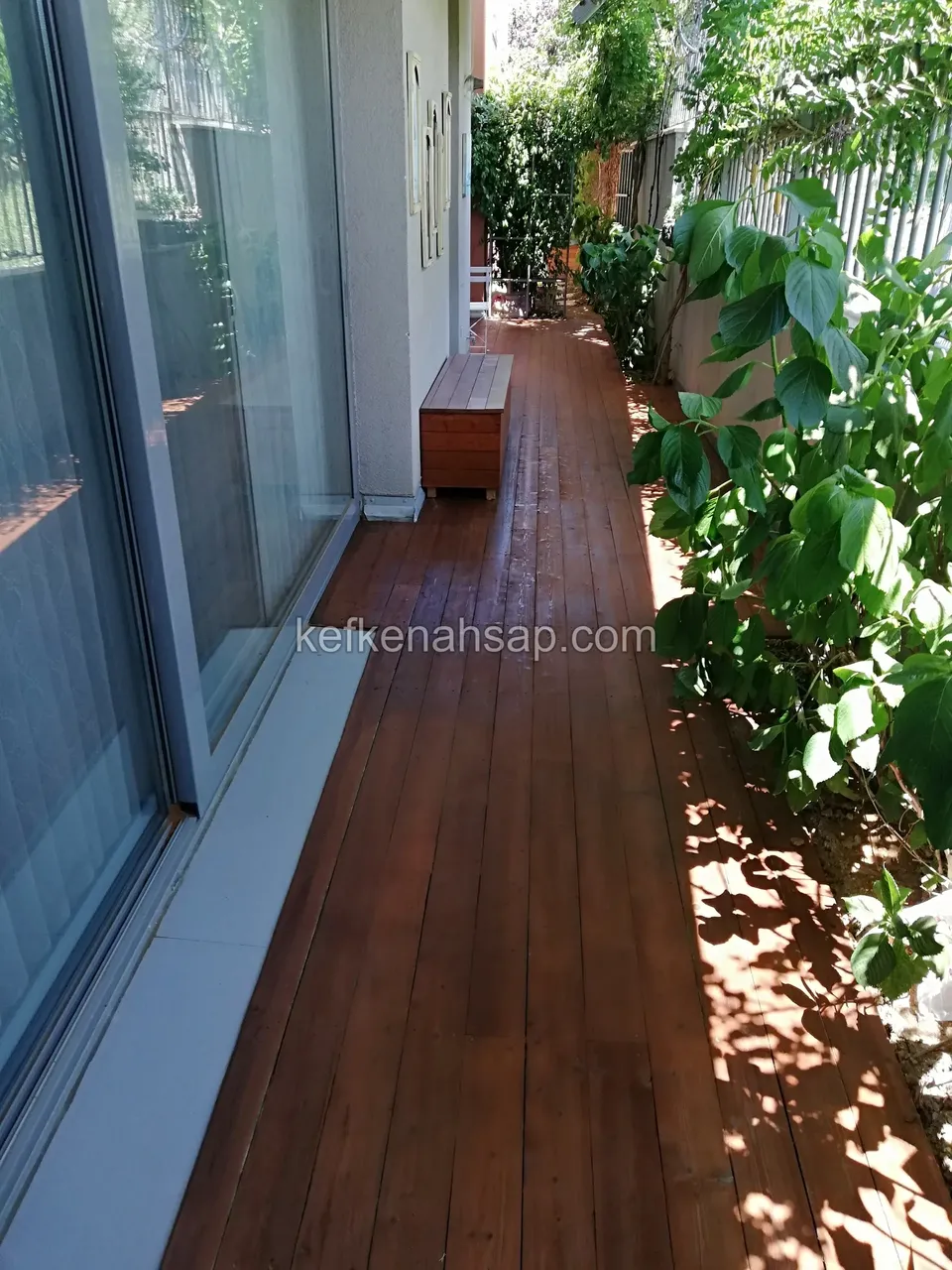 Narrow brown timber deck running along a glass door with plants beside it