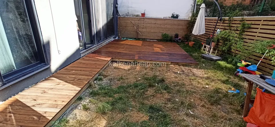 Cam kapı kenarında bahçeye uzanan dar deck yolu; çimen seyrek, yerde oyuncaklar duruyor