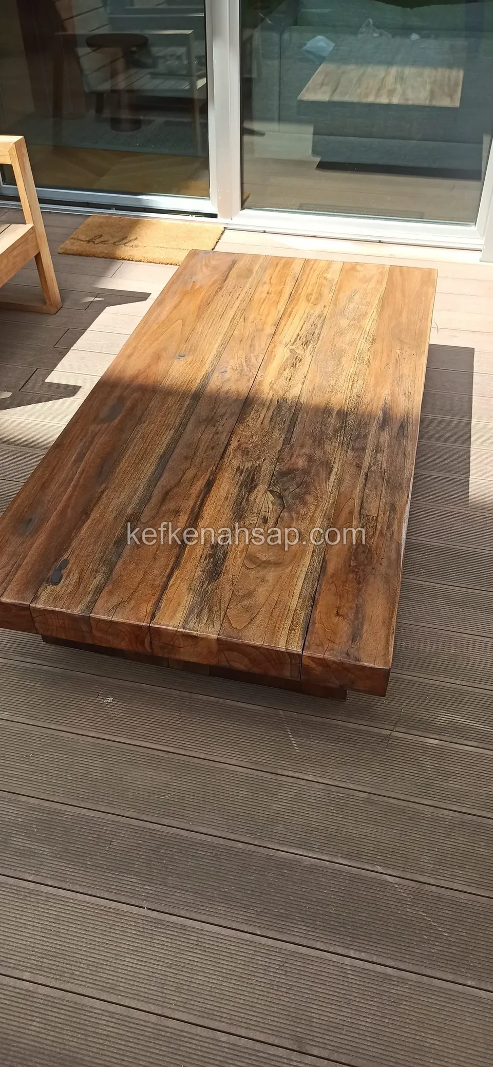On the decking of a terrace with glass doors, a wide, low, solid timber table made of thick, distressed-looking boards