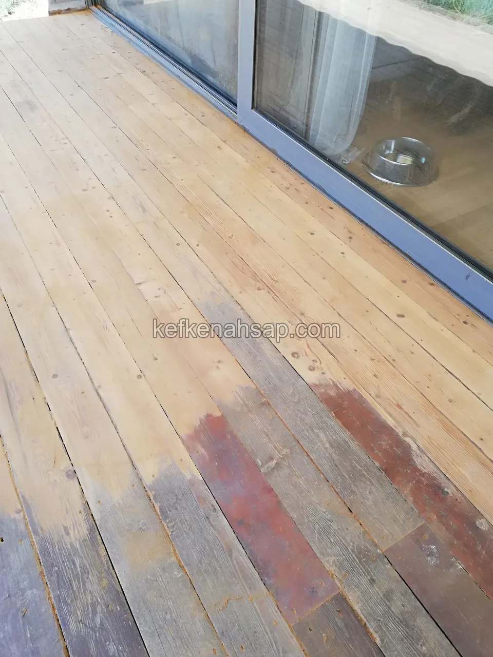 Altes Holzdeck vor einer Glastür, zur Hälfte abgezogen, teilweise ist noch rote Farbe übrig