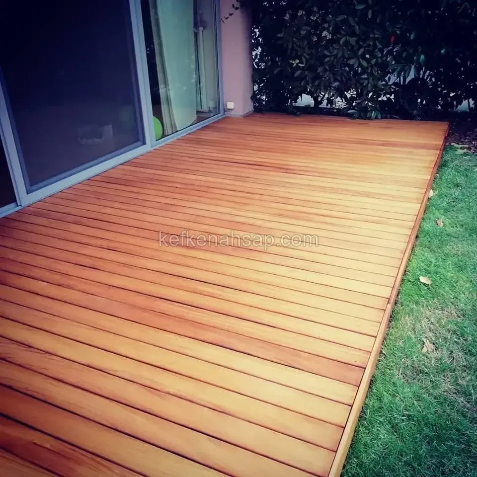 Sürme cam kapıların önüne boyuna döşenmiş, sarıdan turuncuya değişen tonlarda cilalı deck; sağda çimen kenarı
