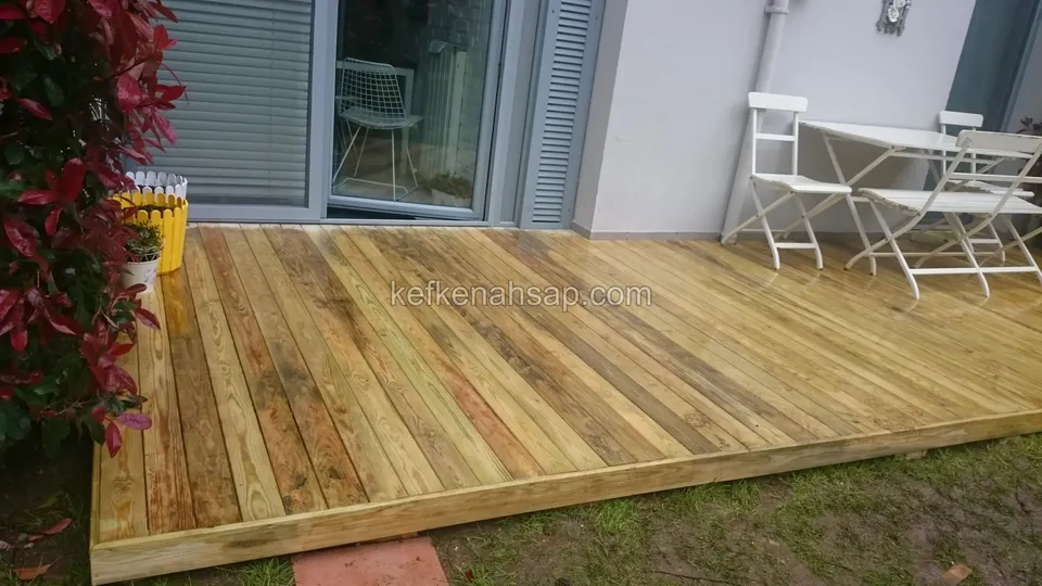 Gri panjurlu cam kapının önüne taze döşenmiş sarımsı emprenyeli çam deck; köşede beyaz katlanır masa ve sandalyeler