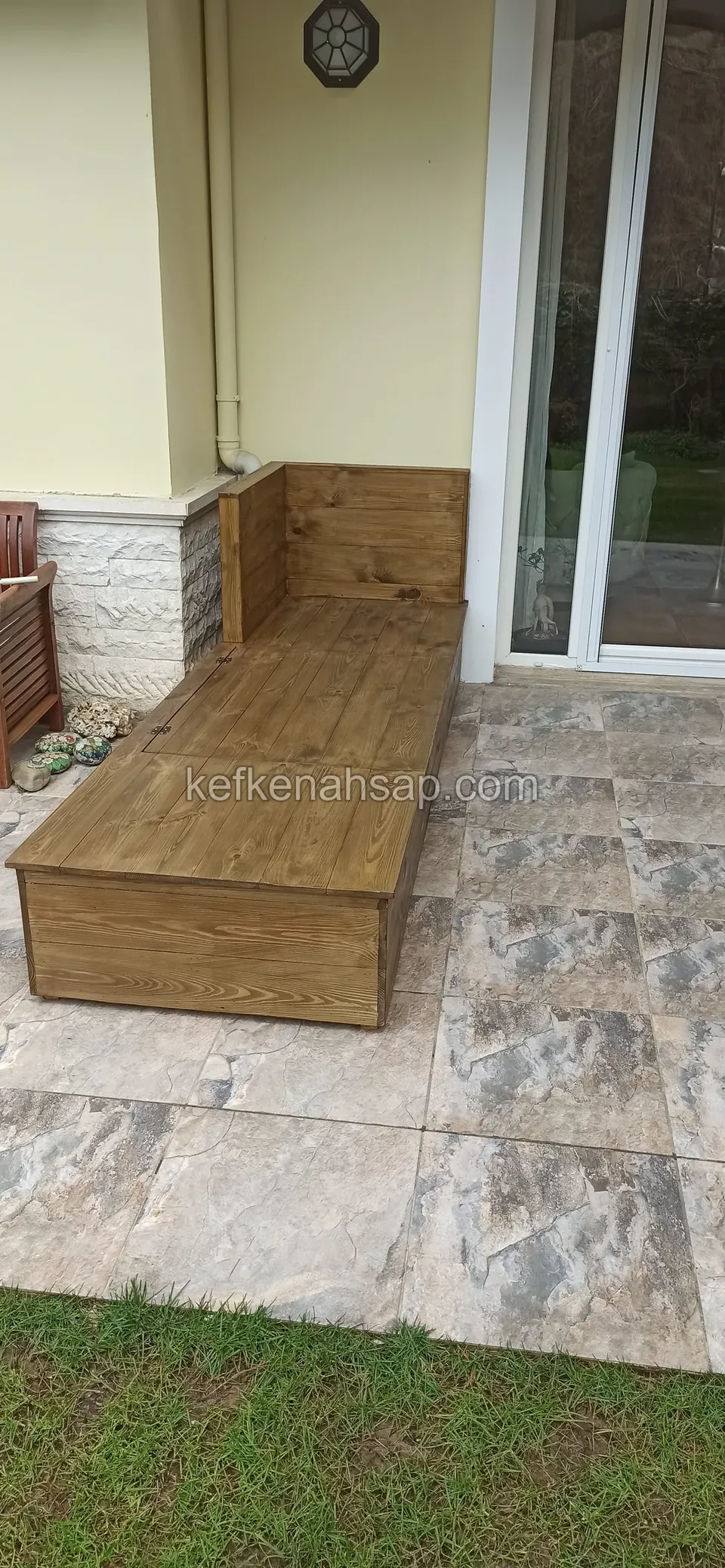 Eine lange Holzliege mit niedriger Lehne auf einer Terrasse mit Fliesen in Steinoptik