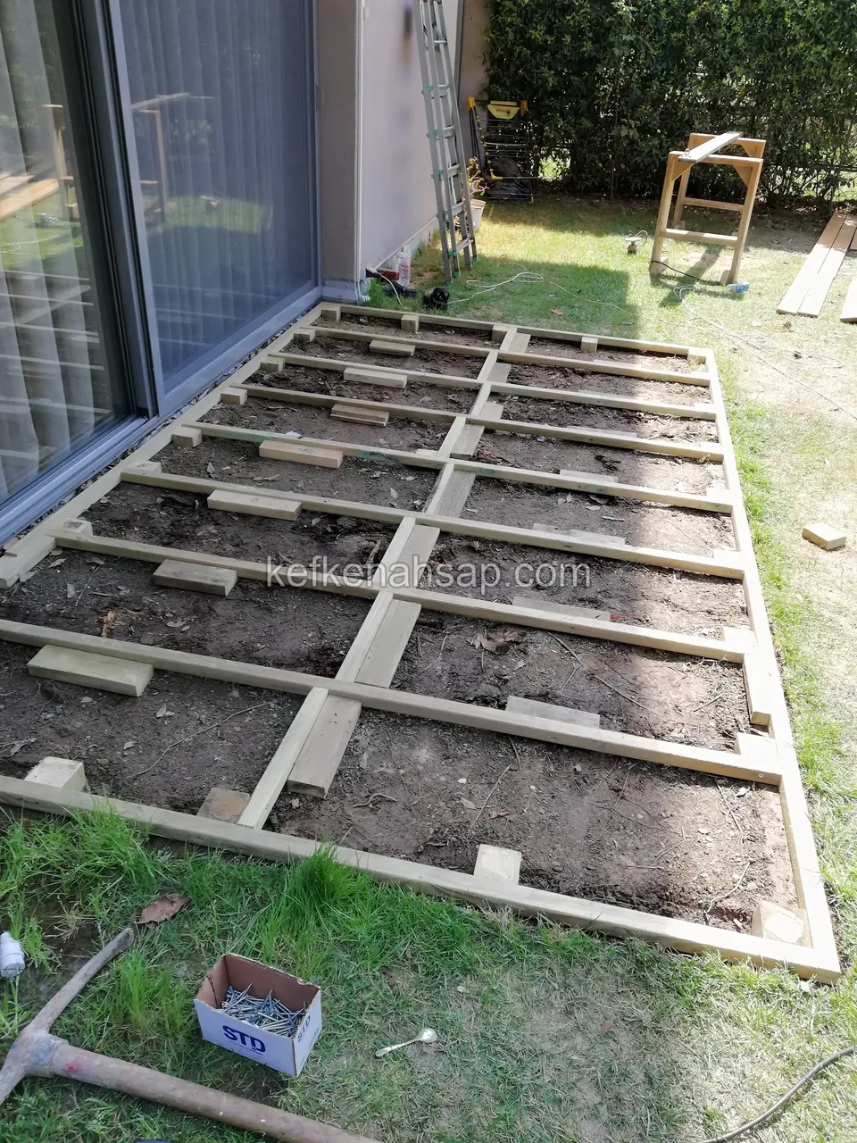 Cam kapı önünde toprak üzerine kurulu ahşap deck alt kiriş çerçevesi; yanda çim ve iş aletleri