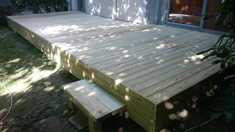Evin cam kapısı önünde yükseltilmiş yeni açık renkli deck ve kenarında ahşap bir basamak; çimde ışık ve gölge benekleri