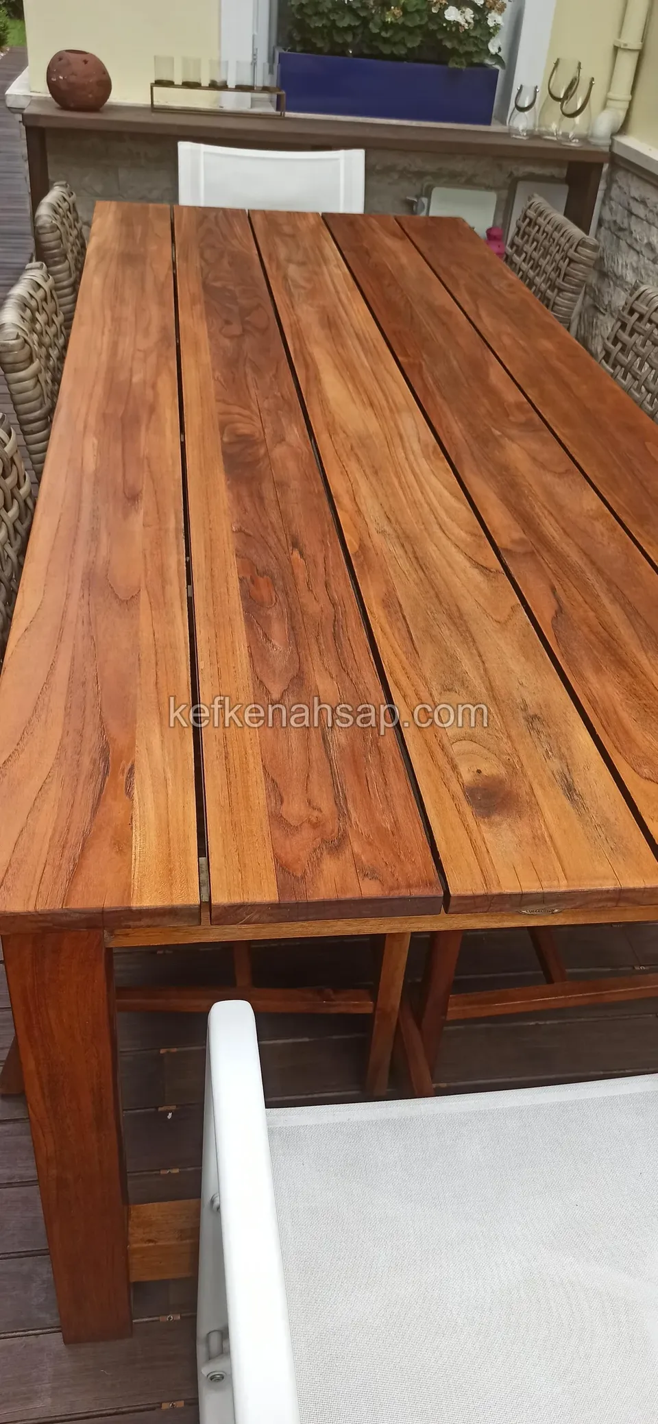 Auf der Terrasse eines Hauses sieht man einen breiten rechteckigen Holztisch aus dicken Bohlen und einen weißen Stuhl am Rand