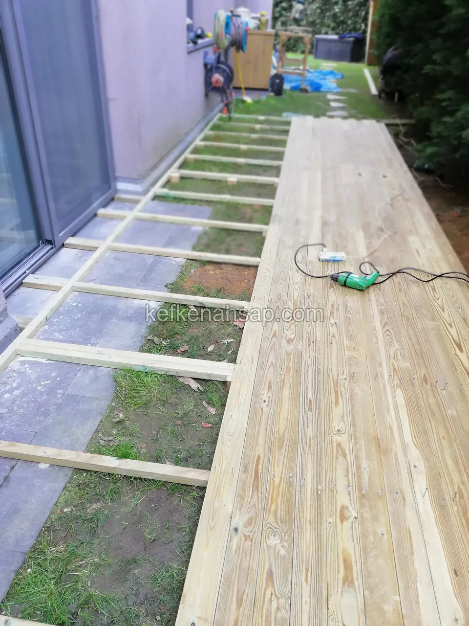 Timber deck partly laid along a narrow side terrace beside a glass door, with a drill on the ground