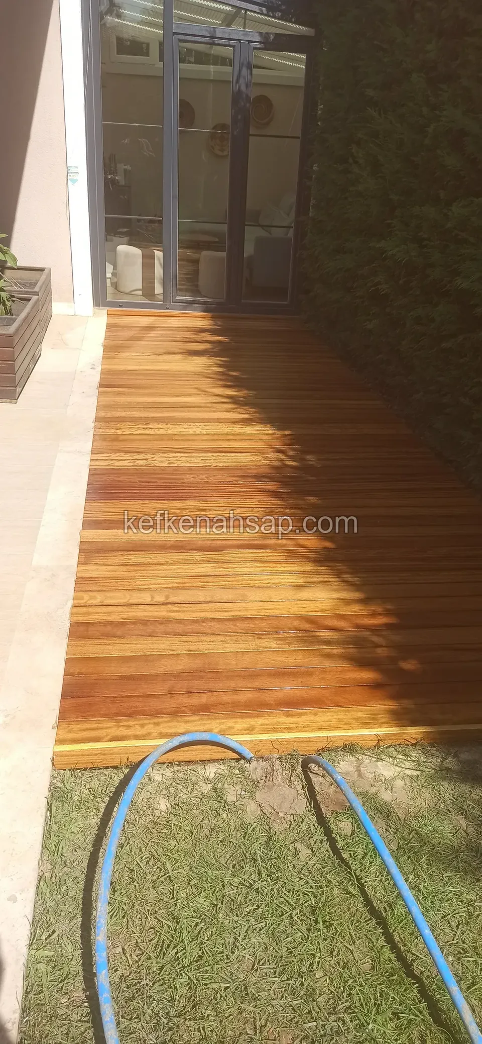 Narrow orange-toned varnished deck running from a glass door out to the lawn, with a blue hose in front
