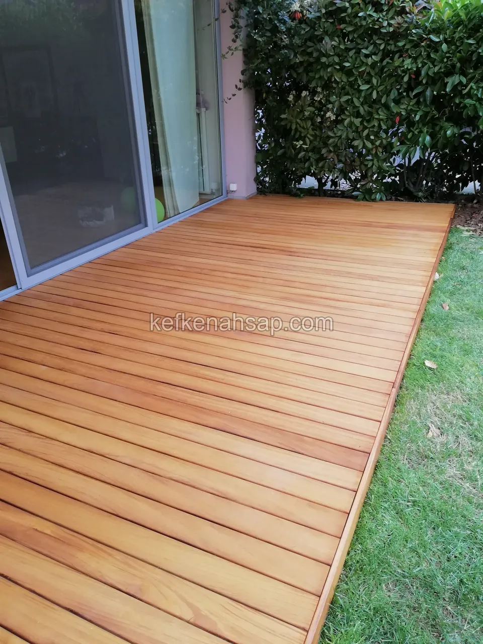 Golden-brown board timber deck in front of glass doors, bordered by lawn on the side