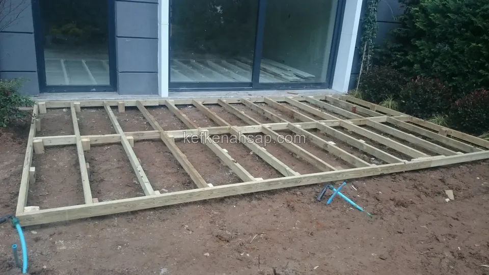 Timber deck substructure set on soil in front of a building with glass doors, joists fixed to a frame
