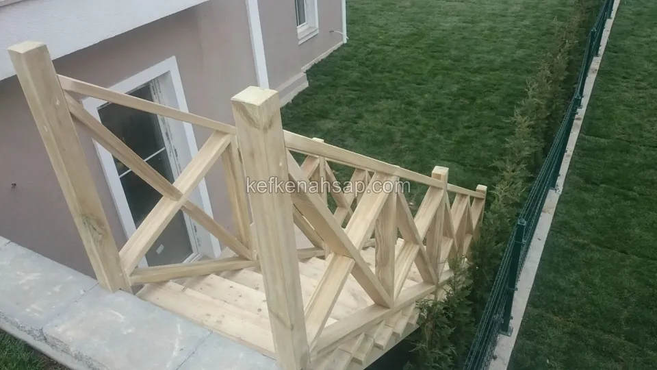 Cam kapılı bodrum girişinin önünde ahşap platform ve çaprazlı korkuluk; yanda çimlik ve tel çit