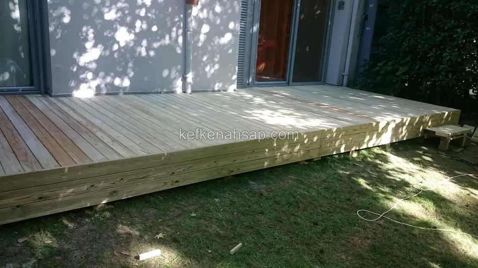 Çimle çevrili, evin cam kapılarının önüne uzanan yükseltilmiş açık renkli deck; kenarında kalın çerçeve tahtası