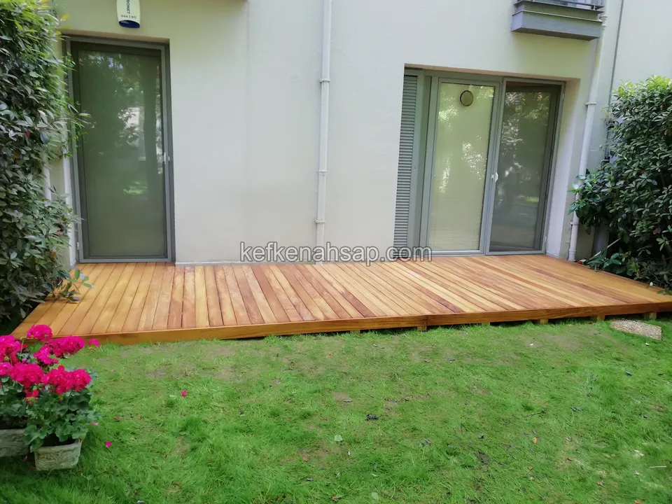 Timber deck bordered by lawn in front of a house with glass doors, with a pink-flowered plant in front
