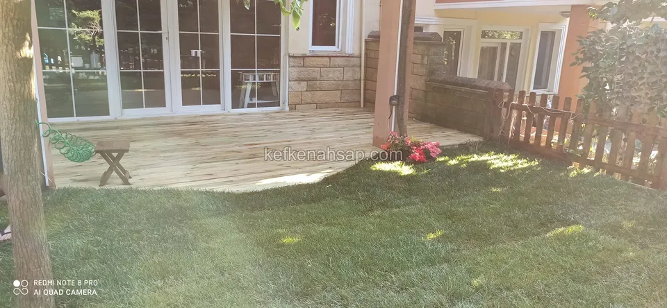 Çimen kenarında, cam kapılı evin önüne serilmiş açık renkli ahşap deck, yanda bir ağaç ve ahşap çit
