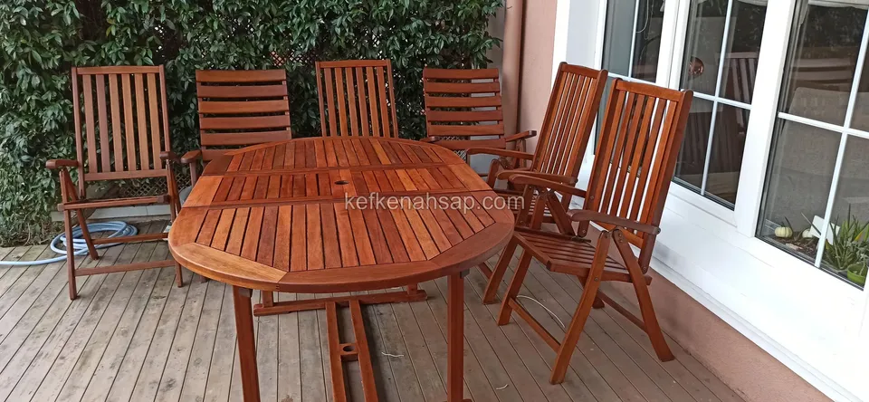 Auf einer Terrasse mit Deck vor grüner Hecke ein ovaler Lattentisch aus Holz mit verstellbaren Holzstühlen